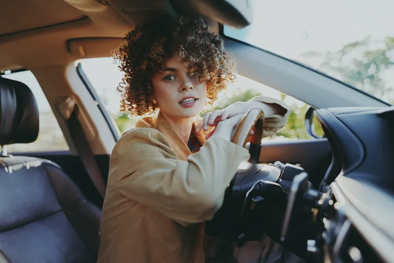 Woman driving a car with a focused gaze, sitting on a leather seat, gripping the steering wheel, showcasing interior details, casual lifestyle shot with retro film color and warm daylight