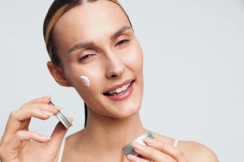A woman applies moisturizer to her face, showcasing a simple skincare routine in a bright studio. A calm, confident expression highlights self care, beauty, and hydration.