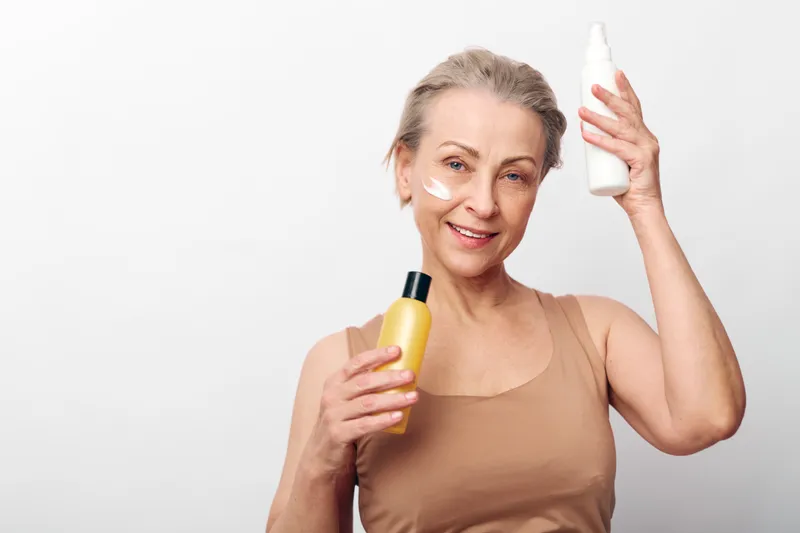 Mature woman with short hair holding skincare bottles applying cream on face in beige tank top. Concept of natural beauty, self-care, and anti-aging routine on white background.