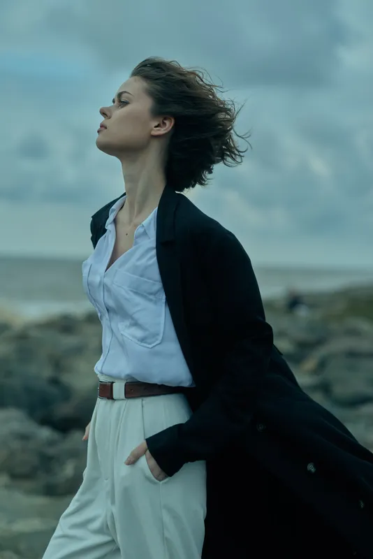 Woman in blazer and white trousers stands on a windy beach, coat fluttering, outdoor professional look with sea backdrop, conveying confidence, freedom and focus in motion