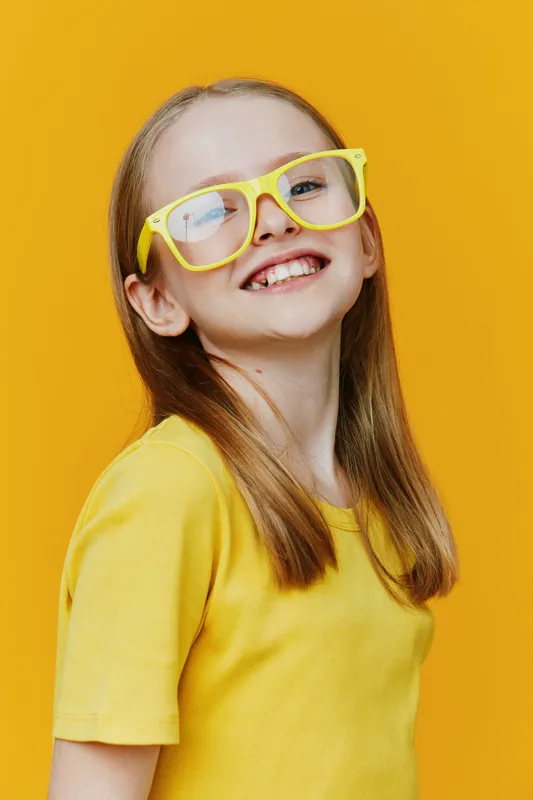 Happy girl wearing yellow glasses and shirt, smiling against a bright yellow background, representing joy and childhood innocence in a vibrant seasonal mood