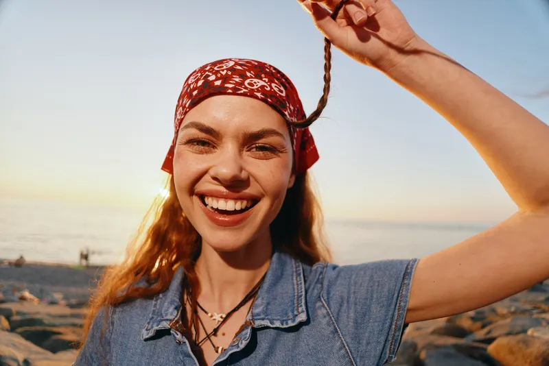 smiling woman with bandana and braided hair wearing denim shirt enjoying sunny beach outdoors with warm light and joyful expression