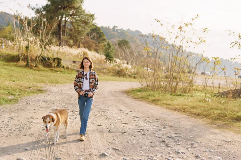Woman walking dog outdoor nature path countryside on a rural dirt road with trees and hills, casual plaid shirt and jeans in a peaceful setting.