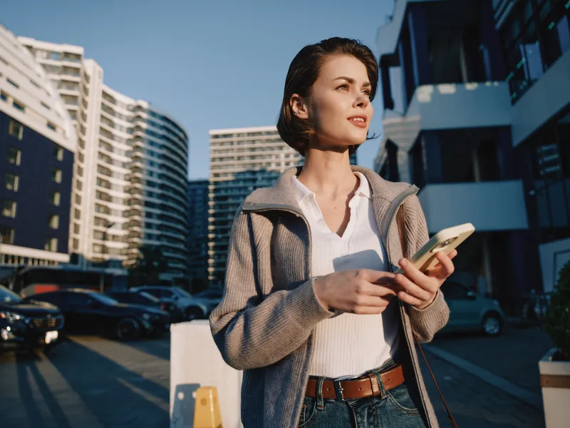 A poised professional woman stands outdoors in a modern urban environment, looking ahead while holding a smartphone. She appears confident and focused, with glass buildings and bright daylight
