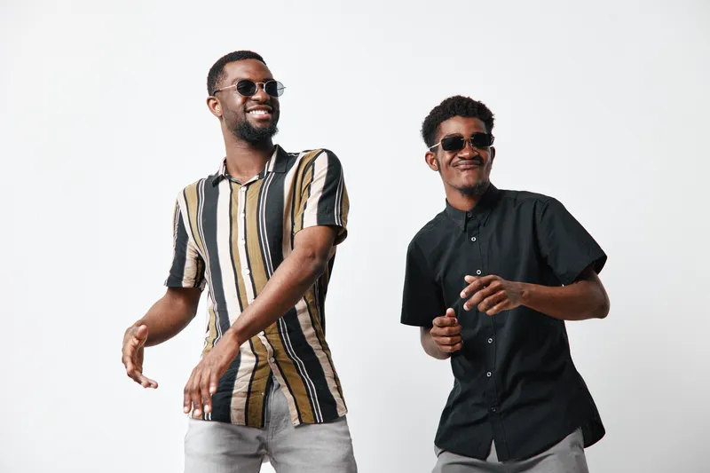 Two young black men with dark skin wearing sunglasses and casual shirts smiling and dancing together on white background for friendship and lifestyle.