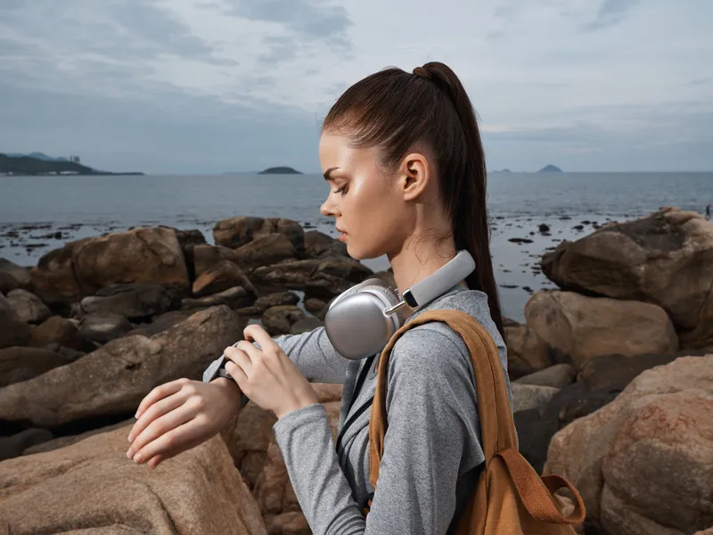 A young woman checking her smartwatch with headphones around her neck, dressed in a casual gray outfit, on a rocky shore with a serene ocean backdrop, reflecting an active lifestyle