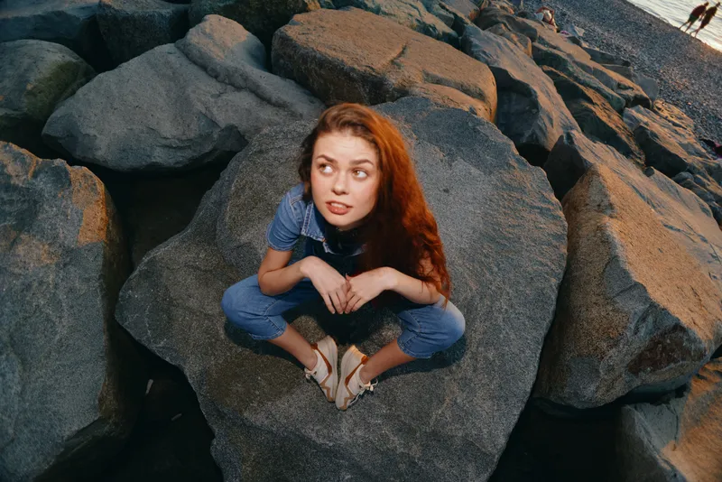 Lifestyle woman in modern boho-western denim street style sitting on rocks with film color effect, capturing relaxed and natural outdoor vibe near water.