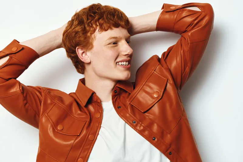 Young redhead man in orange leather jacket smiling and posing with hands behind head on white background. Casual fashion portrait, happy expression and natural look.