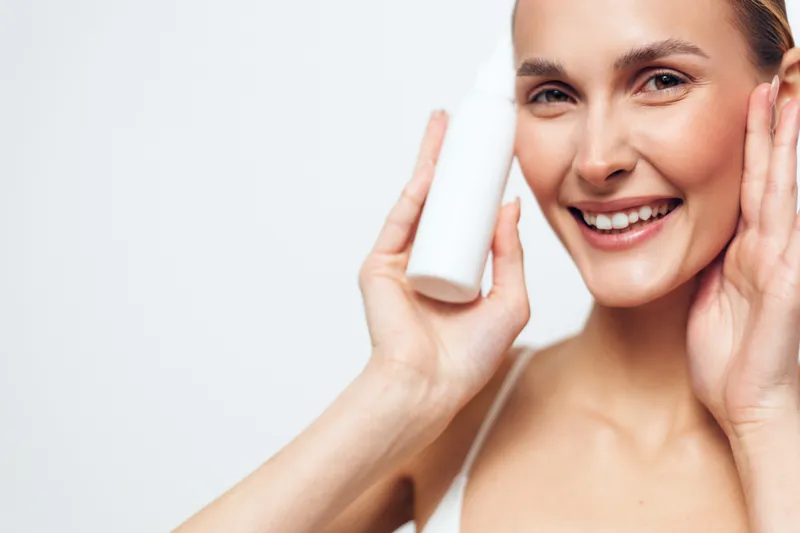A smiling woman holds a white cosmetic bottle near her face with one hand while touching her cheek with the other in a bright studio. Concept for skincare, beauty, and health care.