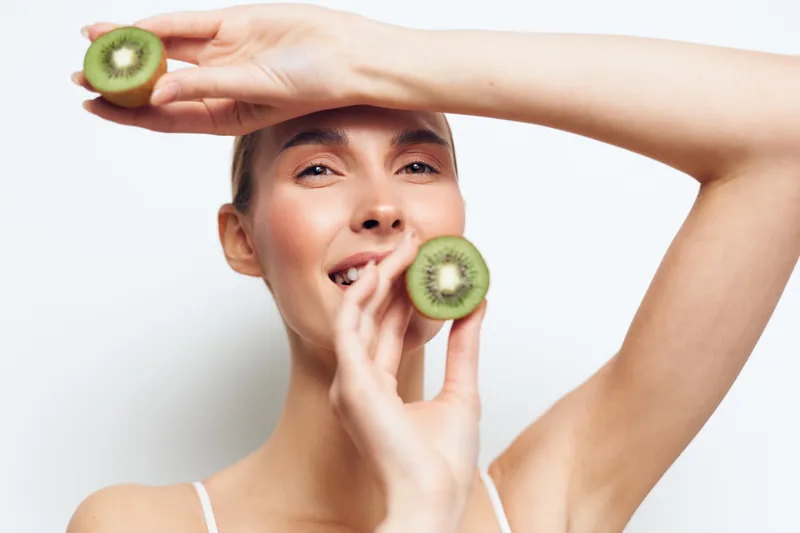 Young woman holding two halves of fresh kiwi fruit near her face smiling with eyes closed on white background. Concept of healthy lifestyle, natural beauty, and wellness.