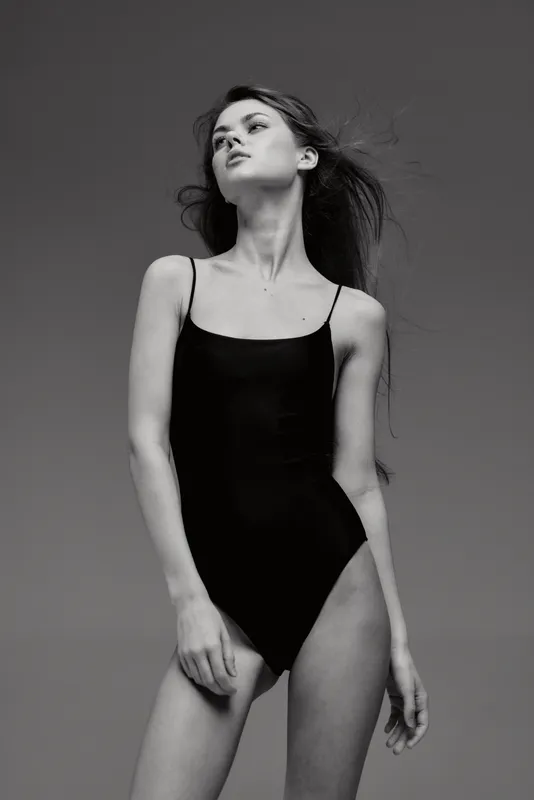 Black and white portrait of a slender woman in a black bodysuit with flowing hair looking upwards in studio.