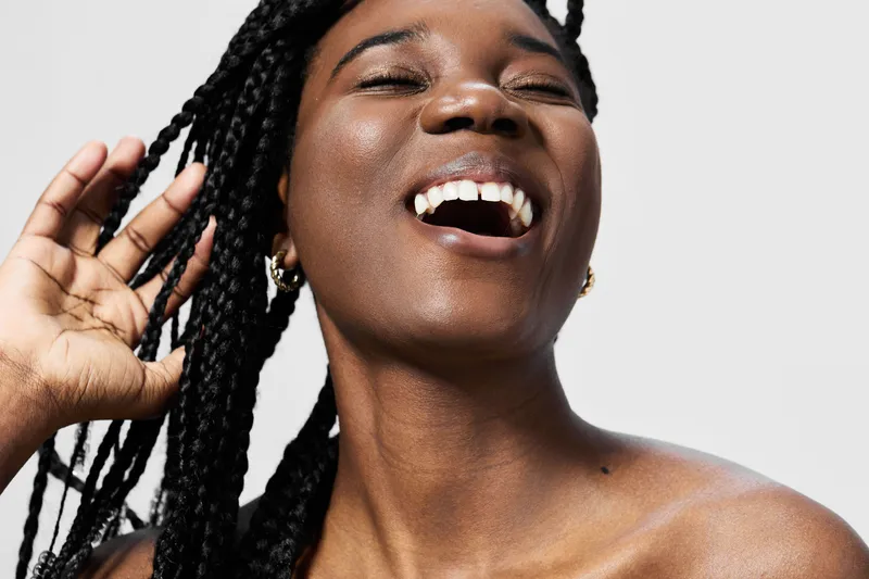 Joyful young woman with braided hair laughing against a minimalistic background, showcasing beauty and confidence in a vibrant, uplifting atmosphere