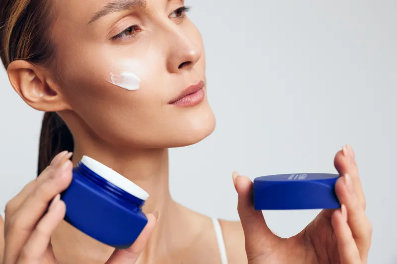 Young woman applying white face cream on cheek holding blue jar and lid on light background. Concept of skincare, beauty routine, self-care, and healthy skin in natural light.
