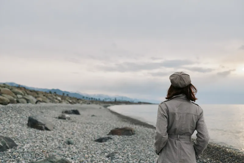 Woman in patterned coat and hat stands on rocky beach looking at calm sea under cloudy sky. Concept of solitude, contemplation, and peaceful nature landscape.