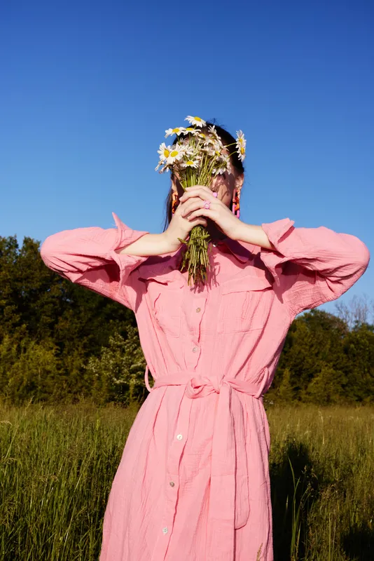 Young woman in a pink dress holding flowers in the field, expressing joy and connection with nature, against a vibrant blue sky backdrop