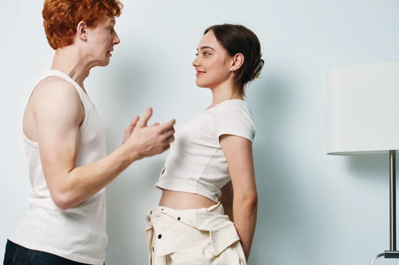 Man and woman standing close in casual clothes indoors, engaging in conversation with focused expressions against light background and modern lamp near wall.