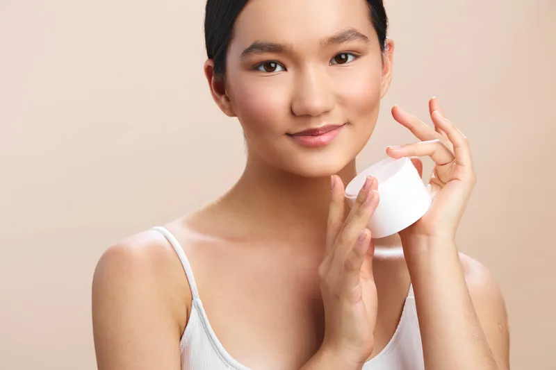 Asian woman applies cream from a jar in a soft studio setting, exuding calm focus and a healthy glow during a skincare routine.
