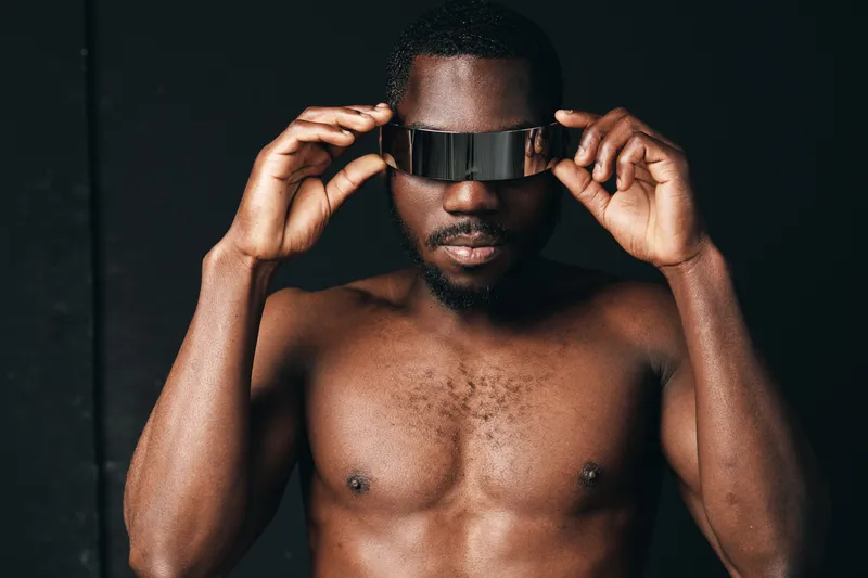 Black man shirtless wearing futuristic visor glasses in creative conceptual visual style with dark background. Portrait of African ethnicity male with modern technology accessory.