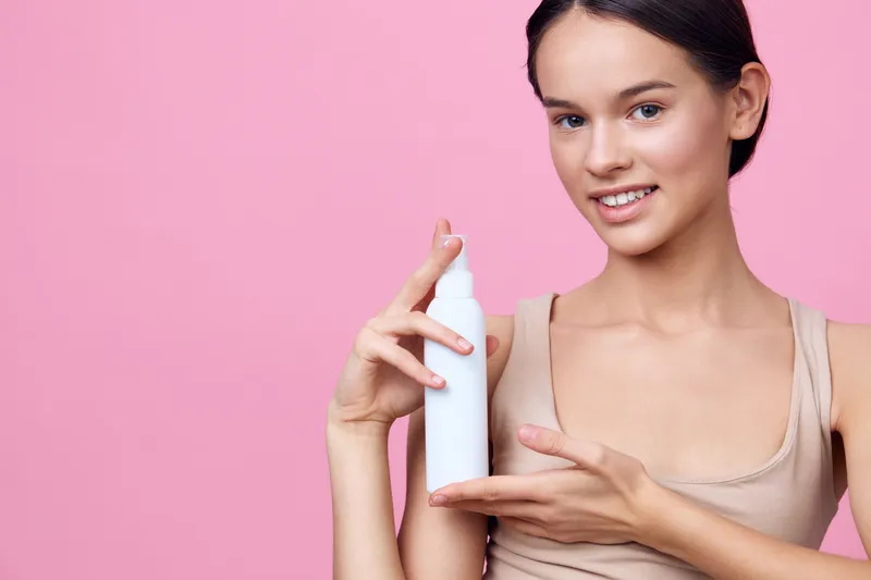 Young woman promoting skincare product on pink background, showcasing healthy beauty with a smile, natural makeup, and soft clothing