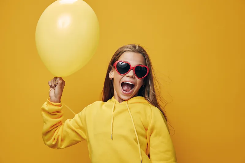 Girl in yellow hoodie with heart shaped sunglasses holding yellow balloon with happy expression on vibrant yellow background for celebration and fun.