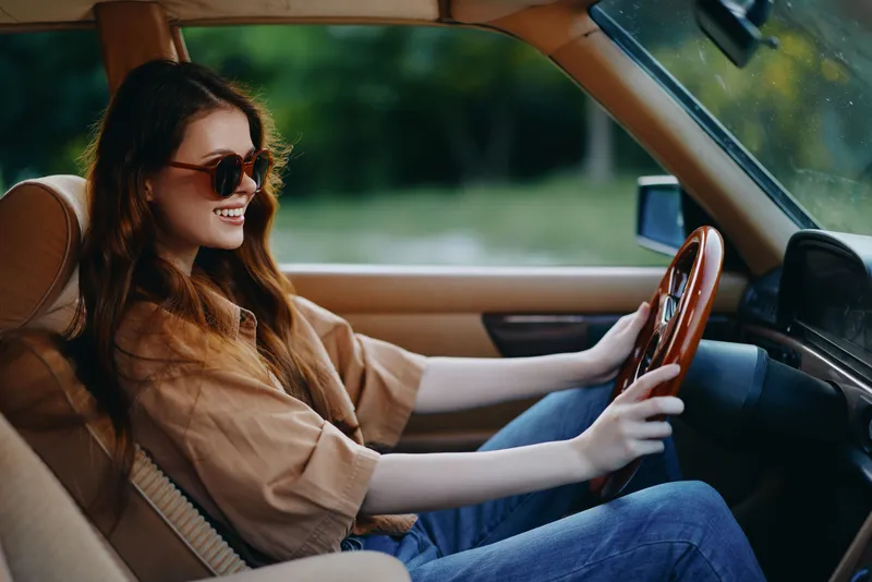Young woman driving a vintage car, smiling joyfully, wearing sunglasses, in a lush green background, conveying a sense of freedom.