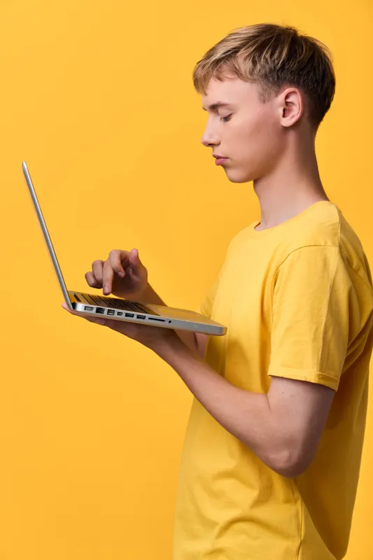 Boy, side view, yellow shirt, using a laptop against a bright yellow backdrop, exploring technology, learning, and digital work in a clean studio scene