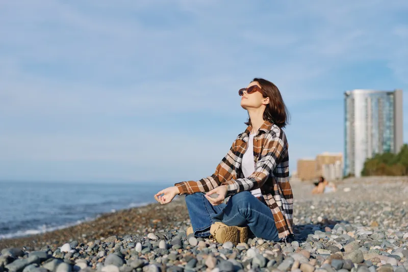 Woman sitting on a pebble beach by the water, wearing a plaid shirt and jeans, looking up at the sky with sunglasses, calm coastal scenery.