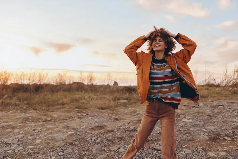 woman lifestyle outdoor sunset portrait casual curly hair joyful walking on rocky path, she stands with hands on head, relaxed smile, warm golden light, field background, nature