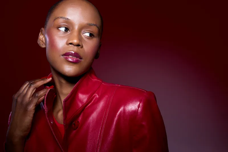 Portrait of a confident black woman with dark skin wearing a red leather jacket against a dark red background. Concept of fashion, beauty, and elegance in studio lighting.