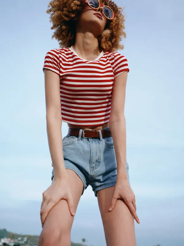 A stylish young woman wearing a red striped tee and high waisted denim shorts poses confidently outdoors under a clear blue sky, exuding carefree summer energy.