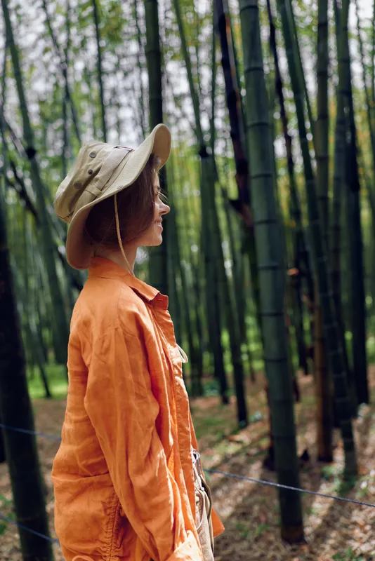 Woman profile wearing a hat and orange jacket in a bamboo forest, side portrait with soft natural light and green vertical trunks, peaceful outdoor scene of travel and contemplation.
