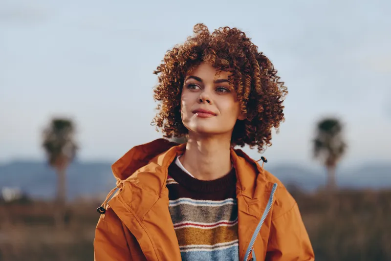 Woman with curly hair wears an orange jacket, knit sweater, stands outdoors in a golden field, calm portrait with mountains, lifestyle concept for fresh air travel, soft bokeh and breeze
