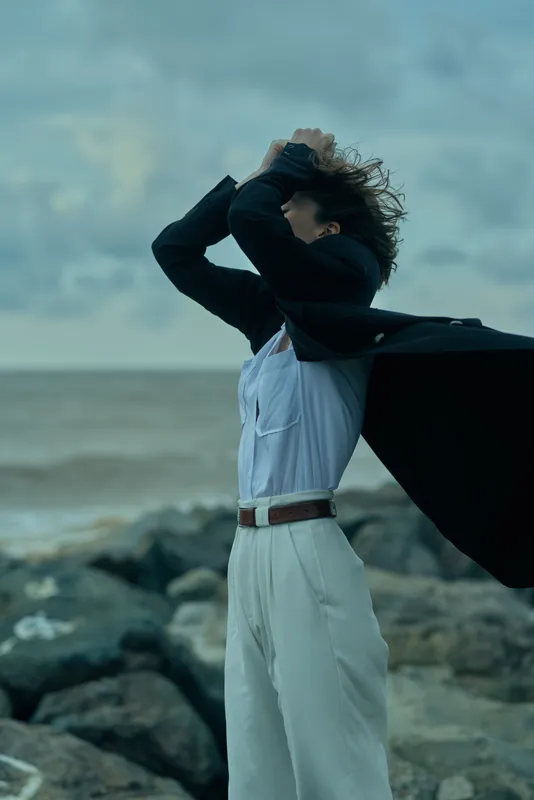 Cape wearer on a windy beach among rocks, arms raised in a dramatic pose, windswept hair, white trousers, ocean horizon behind, cinematic outdoor fashion with energy and freedom in motion