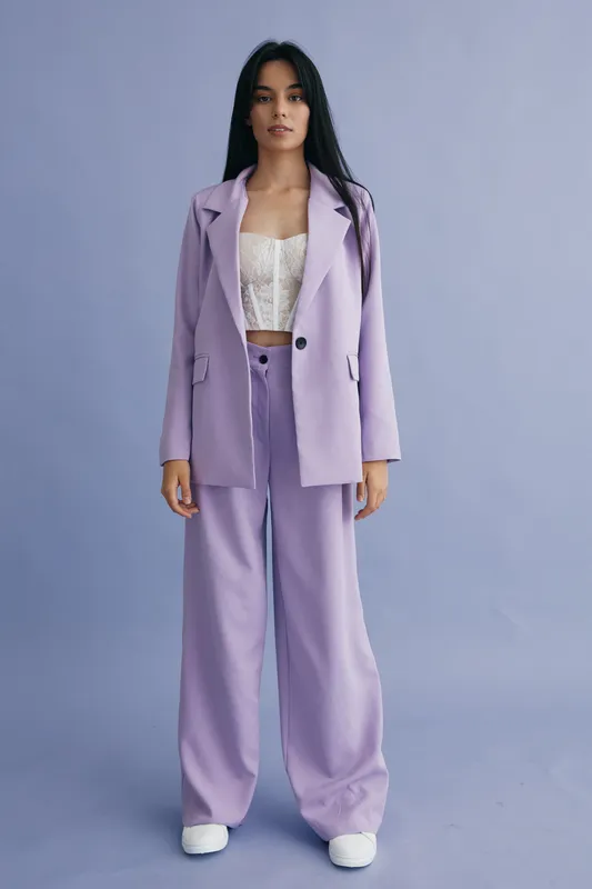 Young woman in lavender pantsuit stands confidently against a soft purple background wearing white lace top and wide-leg trousers in a modern stylish outfit.