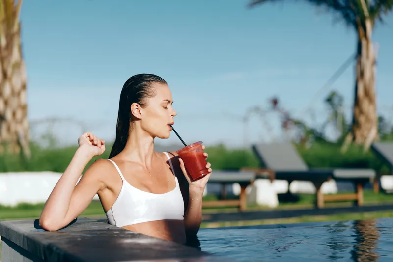 Young woman in a white bikini relaxes in a swimming pool while drinking a red smoothie through a straw on a sunny day with palm trees in the background.