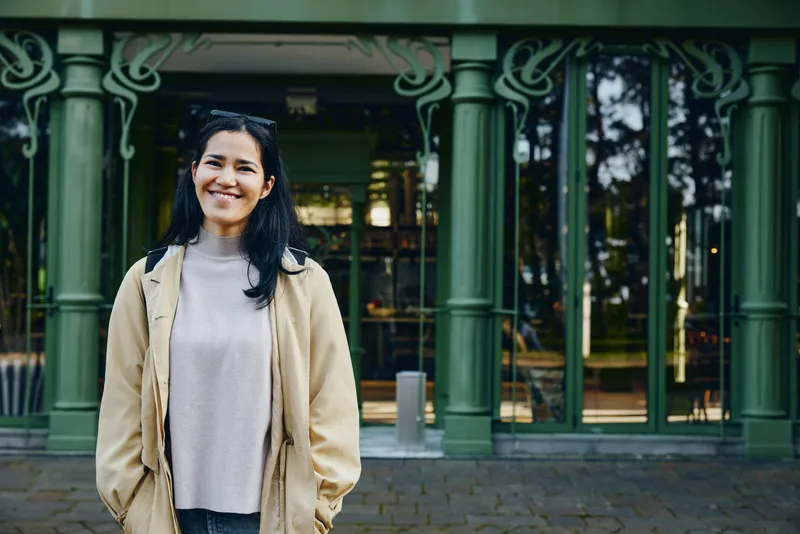 Smiling woman stands outdoors in front of a green ornate building, wearing a beige jacket and gray sweater, candid urban scene with natural light.