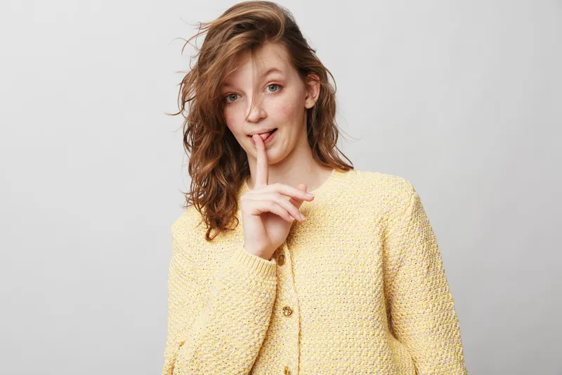 Young woman with curly hair wearing yellow sweater holds finger to lips in a shushing gesture. Studio portrait on grey background with soft natural lighting and casual expression.