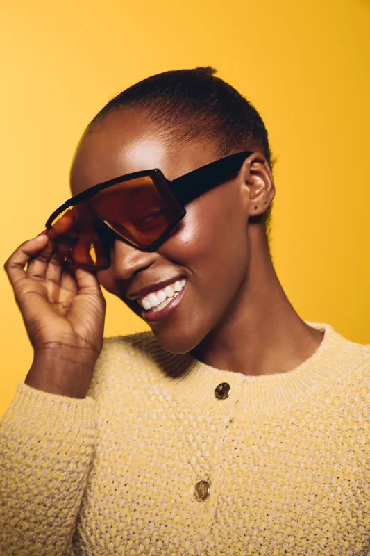 Smiling African woman with dark skin wearing oversized sunglasses and a textured beige sweater poses against a yellow background. Concept for fashion, style, and beauty portrait.