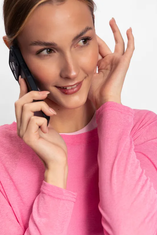 Young woman in pink sweater smiling while talking on smartphone, showing happy expression. Modern communication concept with casual style and positive emotions on white background.