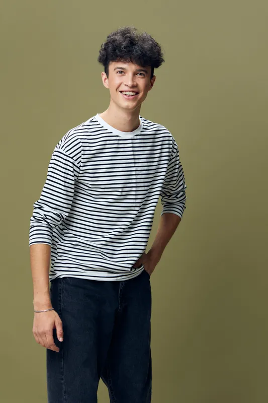 young man smiling wearing striped long sleeve shirt and black pants standing against olive background, casual style, cheerful expression