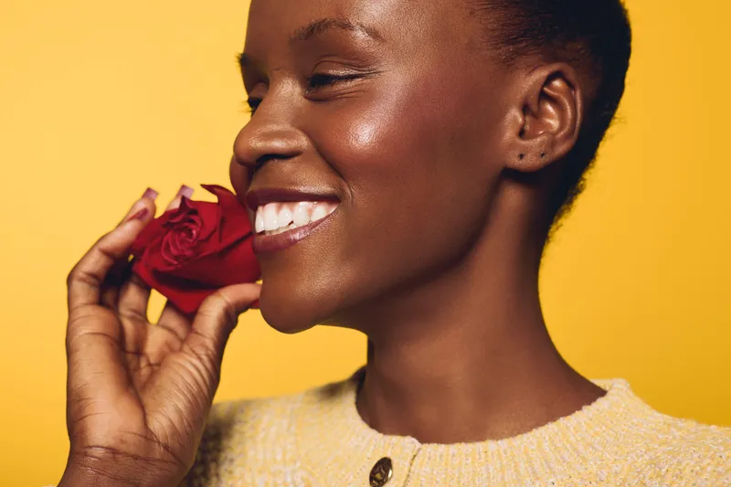 Smiling African woman with dark skin holding red rose near face, eyes closed, wearing yellow sweater. Studio portrait on yellow background. Concept of joy, beauty, and natural skincare.