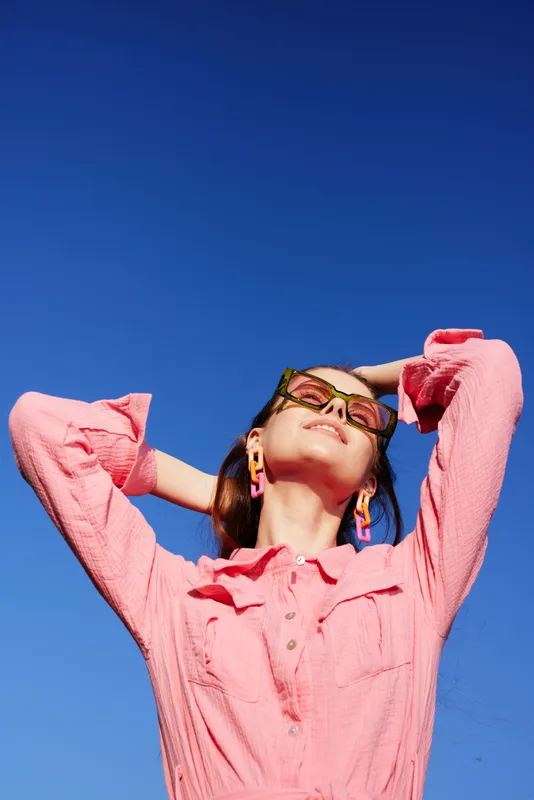 Young woman in pink shirt and sunglasses enjoying the sun against a clear blue sky, evoking feelings of joy and relaxation