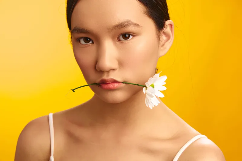 Asian woman smiles softly, with a white flower between her lips, set against a bright yellow backdrop, creating a fresh, elegant portrait with contemporary flair.