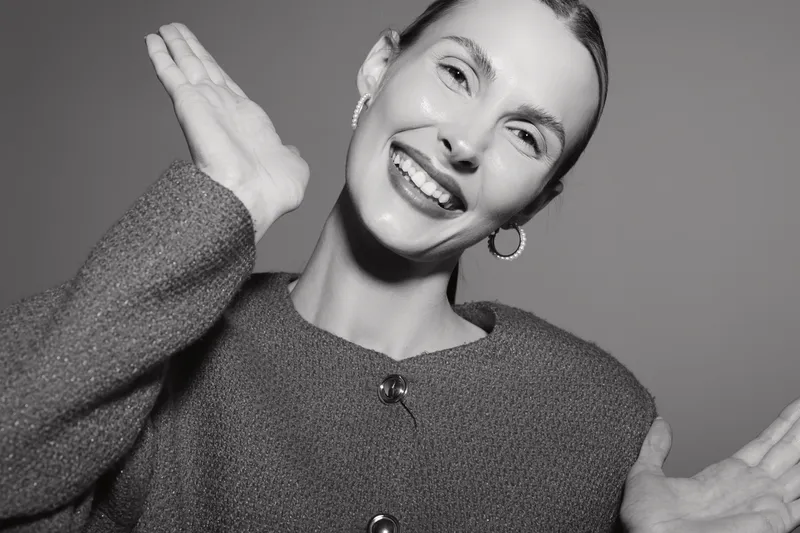 Smiling caucasian woman with light skin wearing a sweater and hoop earrings poses happily in studio with hands raised. Concept for positivity and joyful expression.