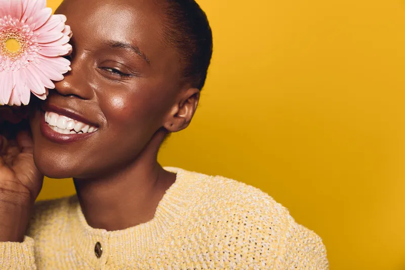 Close-up of a happy African woman with dark skin holding a pink flower near her face. She wears a yellow knitted sweater against a bright yellow background. Concept of beauty and joy.