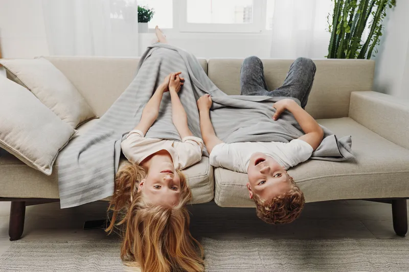 Two happy children lying upside down on a beige sofa covered by a gray blanket in a bright living room. Concept of playful siblings, family time, and cozy home moments.