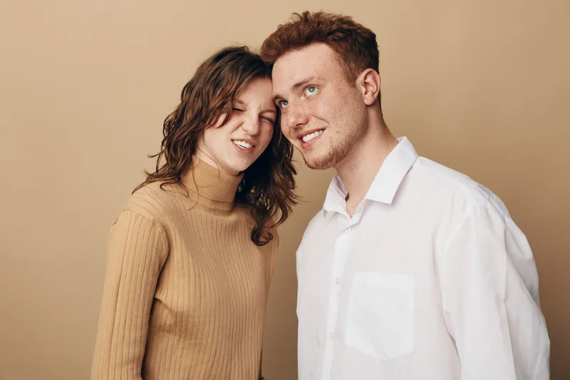 Young man and woman stand close together on beige background. The woman smiles with closed eyes wearing a brown sweater, the man looks sideways in a white shirt. Casual lifestyle concept.