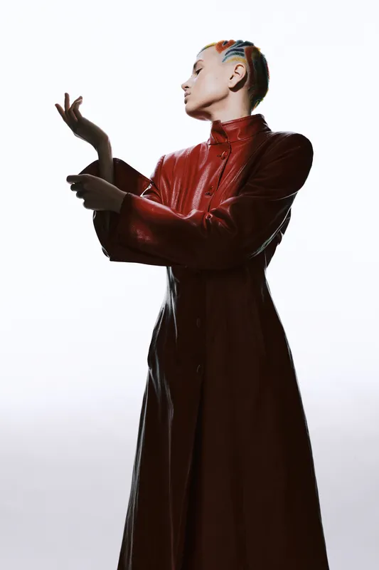 Woman with colorful braided hair wearing a long red leather coat poses gracefully against a clean white background in a high-fashion studio setting.