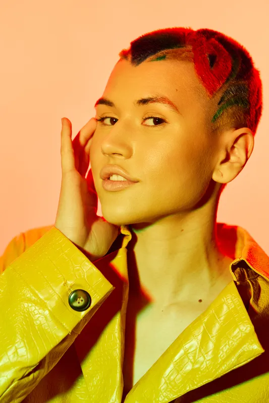 Woman with colorful hair and yellow coat posing confidently against warm peach background, showcasing bold style and modern fashion expression.