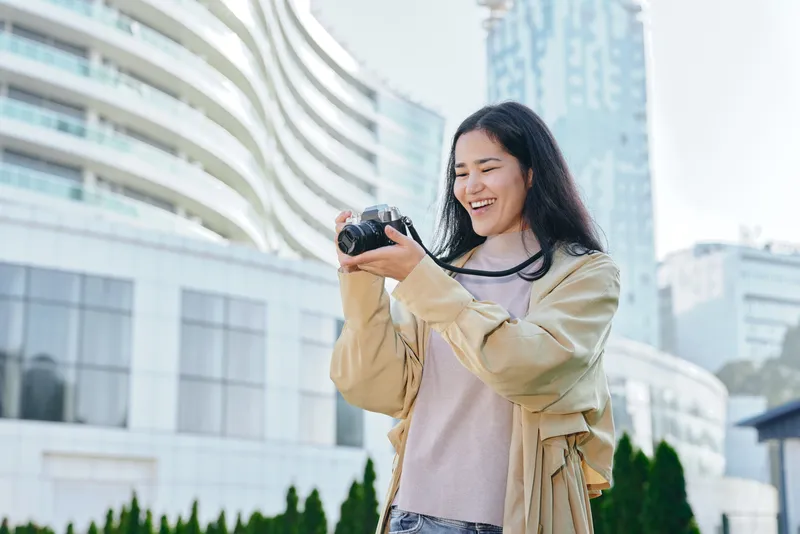 Woman photographer smiling while holding a camera outdoors in an urban area, modern buildings in the background, casual jacket and sweater, candid moment of travel and exploration, street level